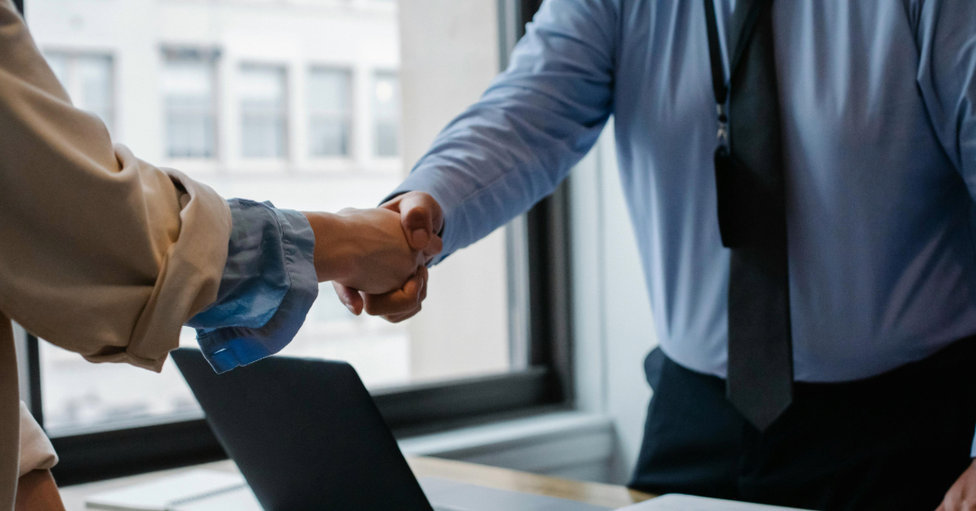 Dos personas se dan la mano ante un laptop, representando relaciones institucionales y colaboración profesional.