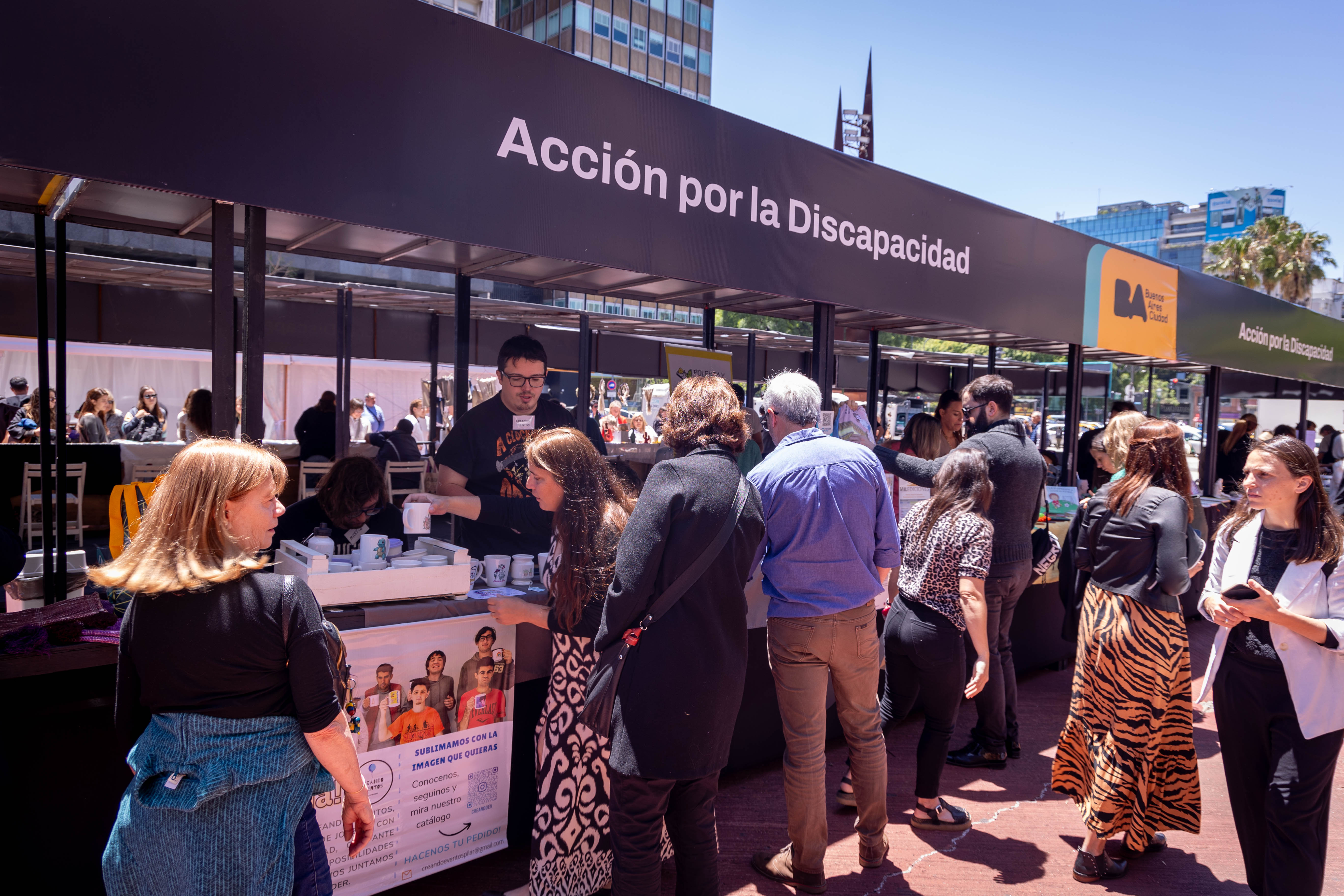 La imagen muestra un evento al aire libre en Buenos Aires, donde hay un gran quiosco decorado con el logo de la ciudad. En el quiosco se celebra la "Acción por la Discapacidad". Varias personas, principalmente mujeres, están haciendo fila para acceder a información o actividades relacionadas con este tema