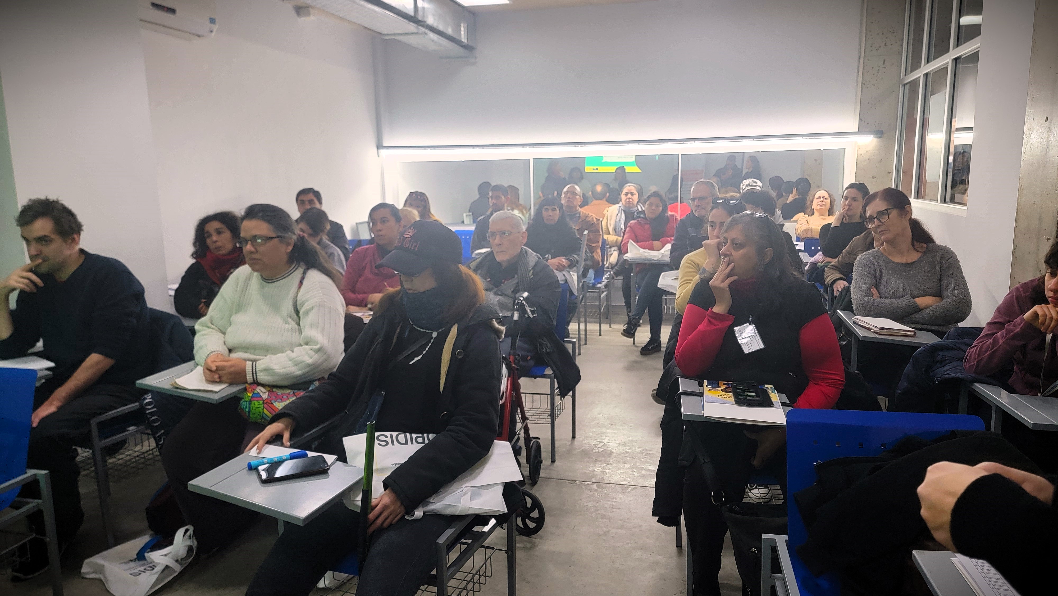 La imagen muestra un aula con un grupo de personas sentadas en escritorios. Hay una variedad de asistentes, tanto hombres como mujeres, de diferentes edades. La mayoría parece estar prestando atención, con algunos mirando hacia el frente y otros revisando sus notas o dispositivos. La iluminación es brillante y el ambiente parece ser de aprendizaje. Al fondo, se puede ver una ventana grande que permite la entrada de luz natural. Algunos asistentes llevan materiales de estudio, como libros y cuadernos. La disposición de los escritorios sugiere un formato de clase o taller.