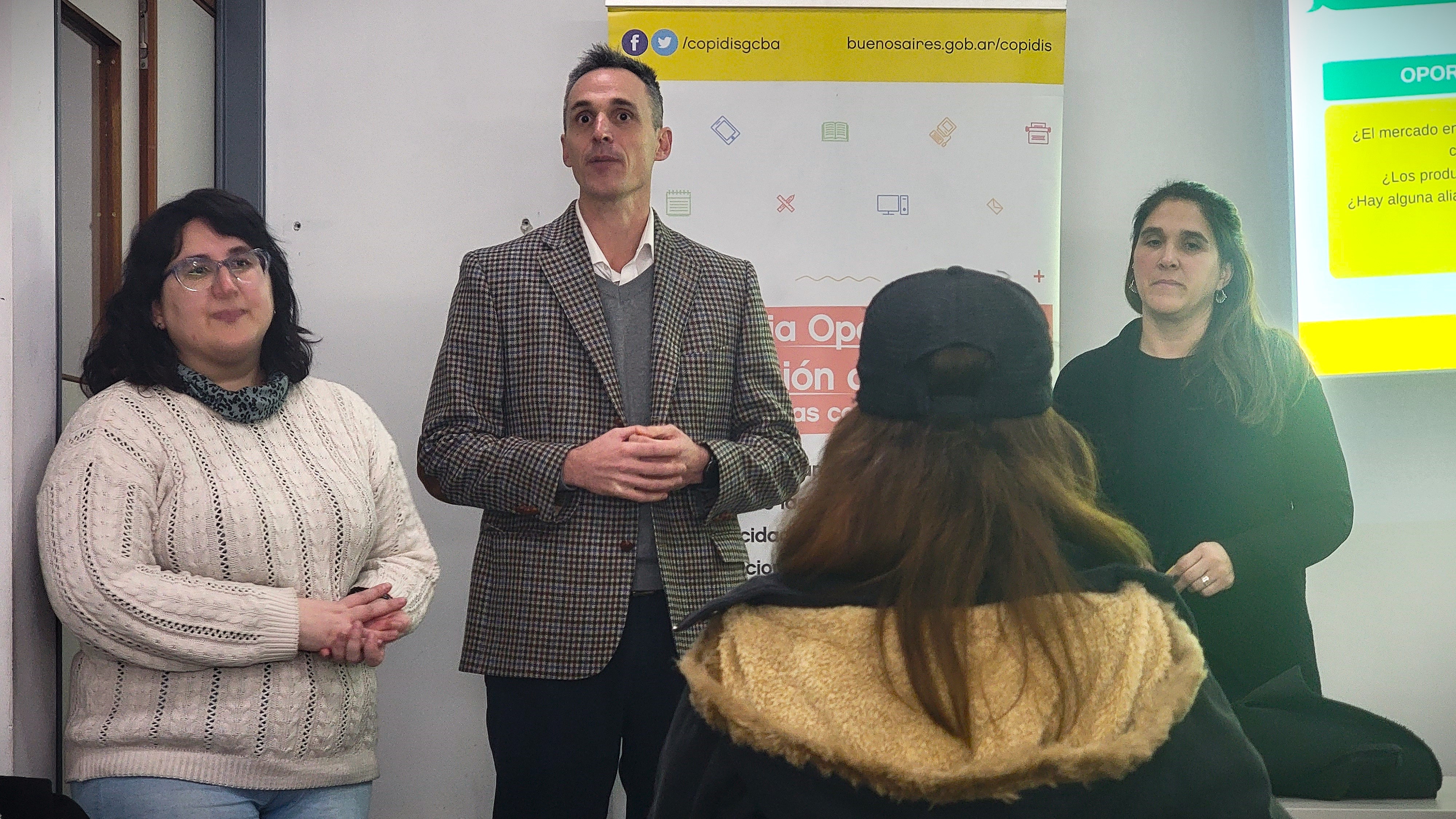 La imagen muestra a tres personas en un entorno de presentación, La capacitadora, Pedro Crespi y una integrante del equipo del Registro Laboral de COPIDIS, en el aula o sala de conferencias. En el centro, Pedro Crespi con un traje de cuadros está de pie, hablando y parece estar explicando algo a la audiencia. A su izquierda, hay una mujer con cabello rizado y gafas, que escucha atentamente. A la derecha, otra mujer con cabello largo y lacio también está prestando atención. Al fondo, se puede ver un cartel con información. La atmósfera parece ser de interés y participación.