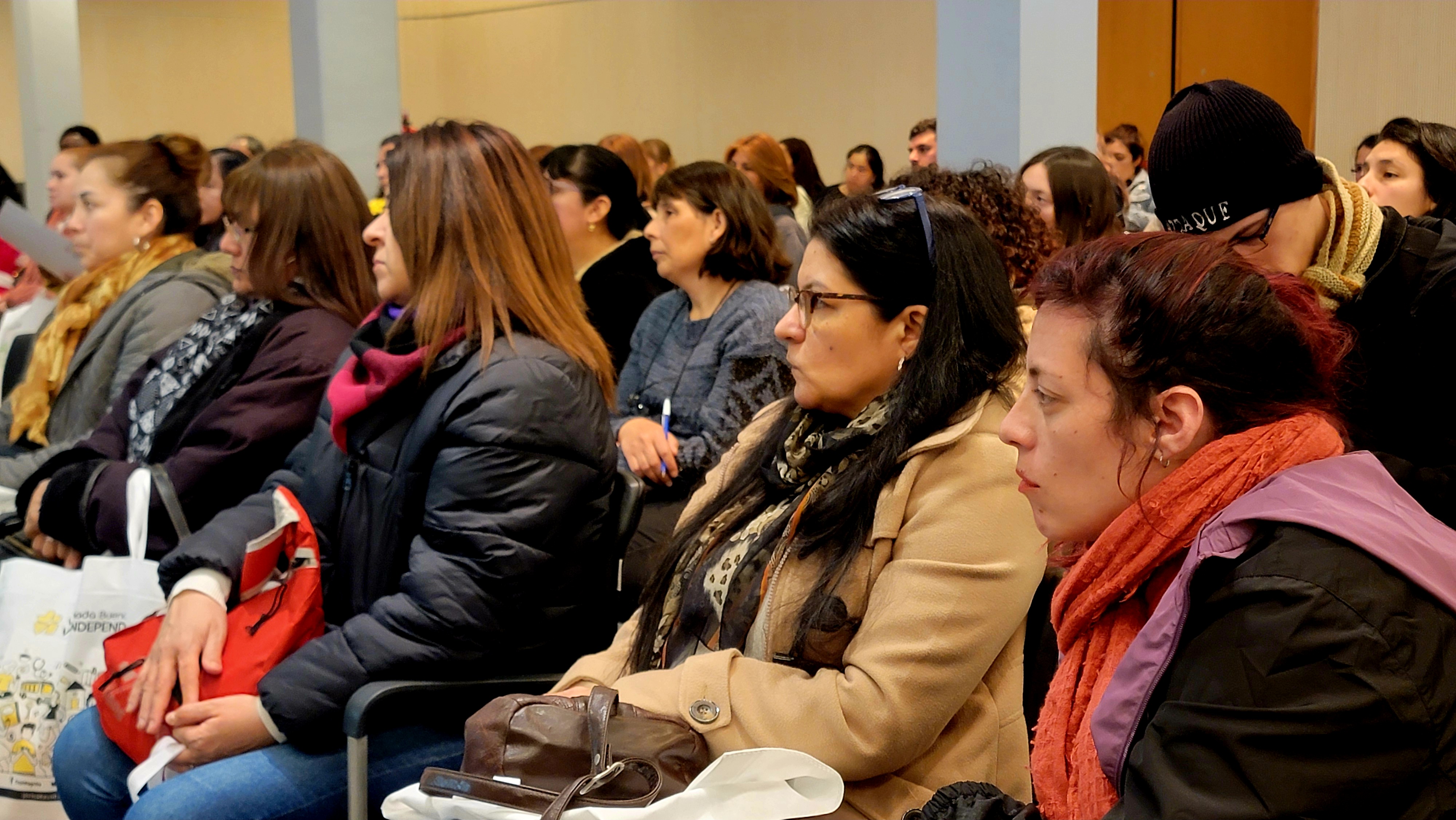 La imagen muestra a un grupo de mujeres sentadas en un auditorio, prestando atención a la presentación o charla. La mayoría de ellas lleva ropa de abrigo, como chaquetas y bufandas, lo que sugiere que el ambiente es fresco. Las expresiones en sus rostros son serias y concentradas, indicando que están involucradas en la actividad. Algunas tienen bolsas o mochilas a su lado. En el fondo, se pueden ver más personas, lo que sugiere una buena asistencia al evento. La disposición de las sillas y la atención del público reflejan un ambiente de aprendizaje o discusión.