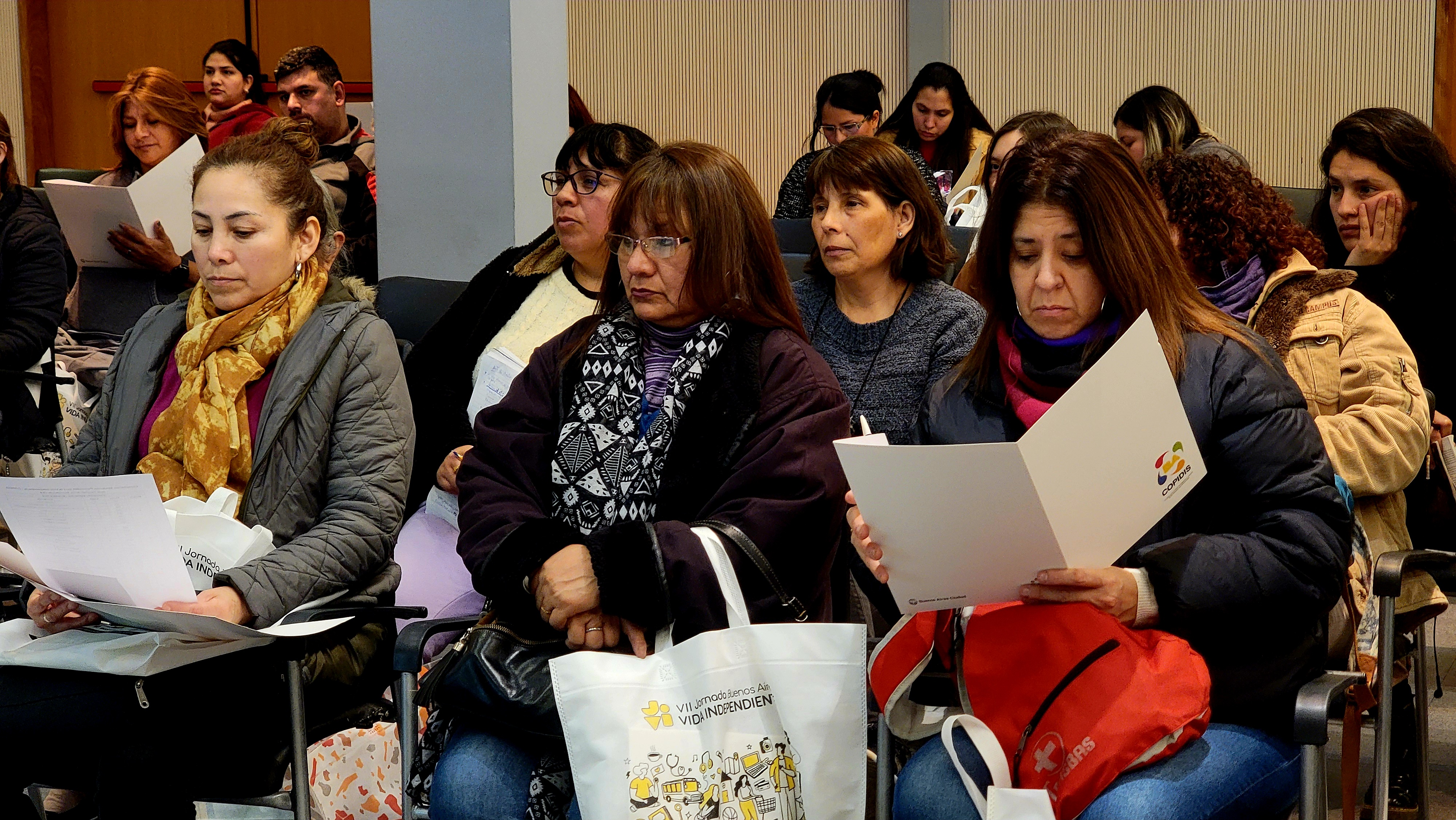 La imagen muestra a un grupo de mujeres sentadas en un auditorio, concentradas en la lectura de documentos que tienen en sus manos. La mayoría de ellas viste ropa de abrigo, como chaquetas y bufandas, lo que sugiere que el ambiente es fresco. Algunas tienen bolsas a su lado, con materiales relacionados con el evento. Las expresiones en sus rostros son serias y atentas, indicando que están involucradas en la capacitación o presentación. En el fondo, se pueden ver más asistentes, lo que sugiere una buena participación en la actividad. La atmósfera refleja un ambiente de aprendizaje y compromiso.