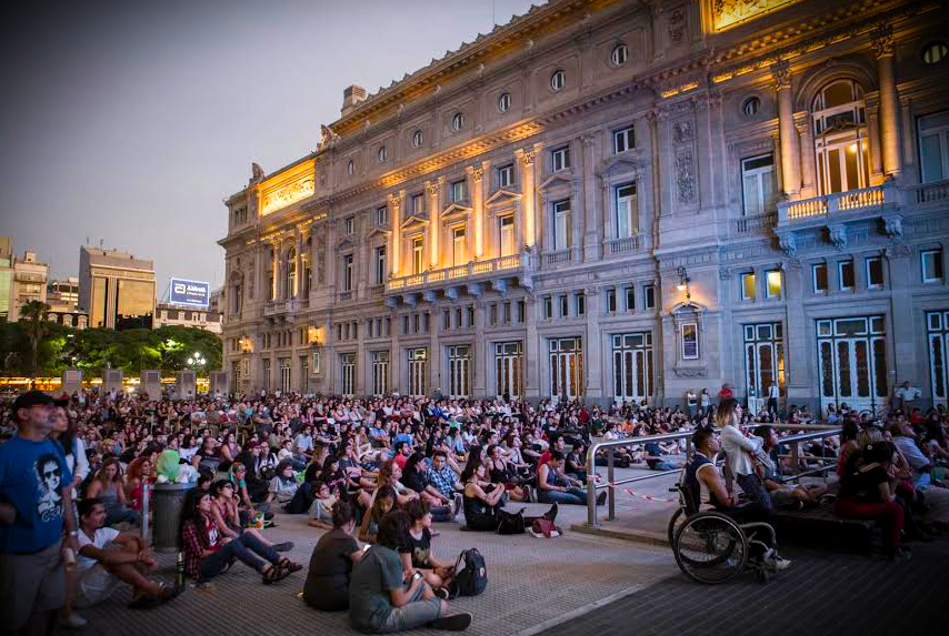 La imagen muestra una gran multitud reunida en plaza vaticano, frente al majestuoso Teatro Colón iluminado por la tarde. El público está sentado en el suelo, con rostros que reflejan interés y expectación, lo que sugiere que están esperando algún tipo de evento o espectáculo.