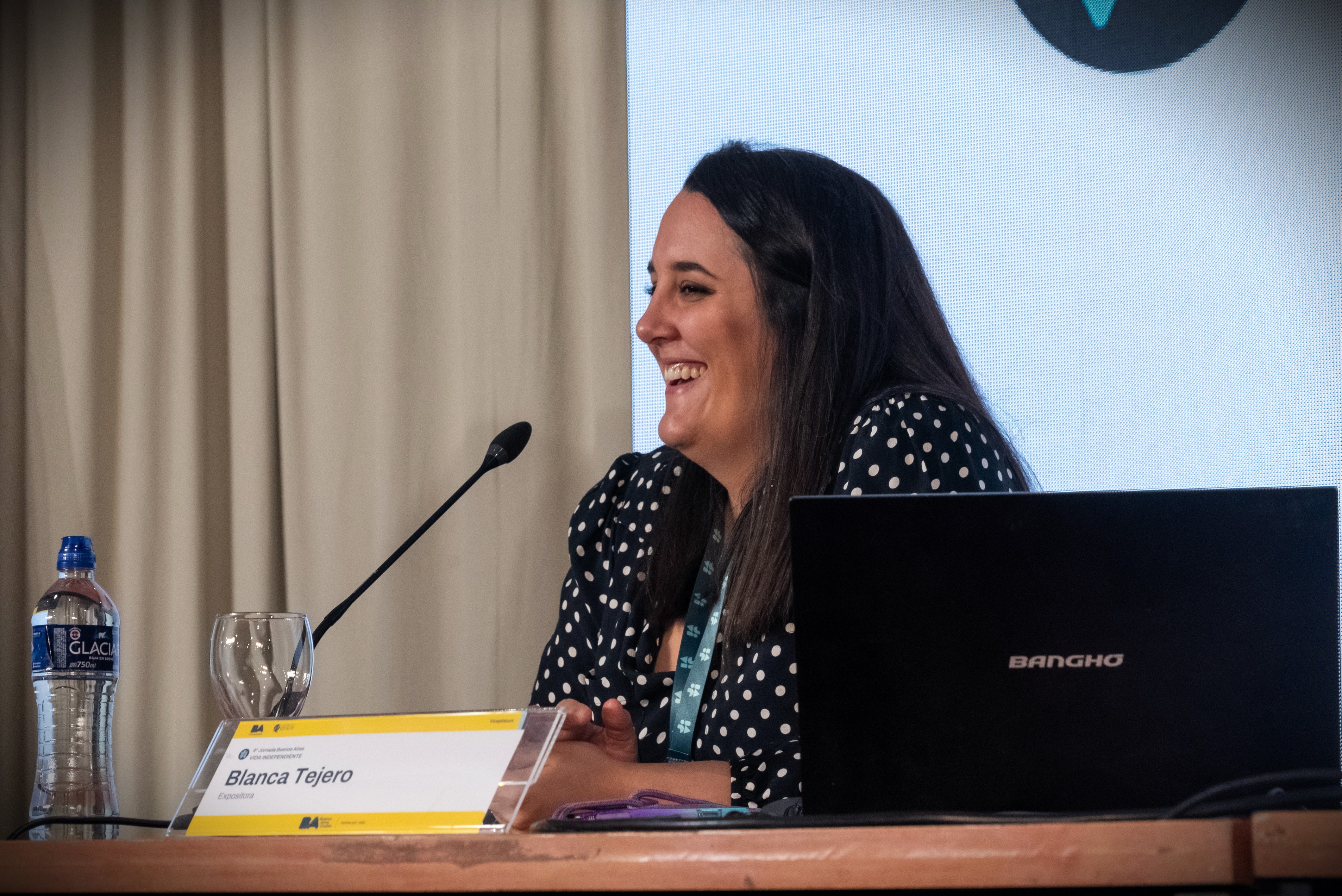 En la imagen se puede ver a Blanca Tejero, que está participando en un evento. Ella tiene el cabello oscuro y lleva una blusa de lunares blancos sobre fondo negro. Su expresión es alegre y sonriente, lo que sugiere que está disfrutando del momento.  Está sentada en una mesa con un portátil frente a ella y una botella de agua al lado. También se observa un micrófono, indicando que está participando en una  presentación. El fondo es de color claro, y hay una pantalla detrás de ella que podría estar mostrando información relevante al evento. En general, la atmósfera parece ser de interacción y diálogo.