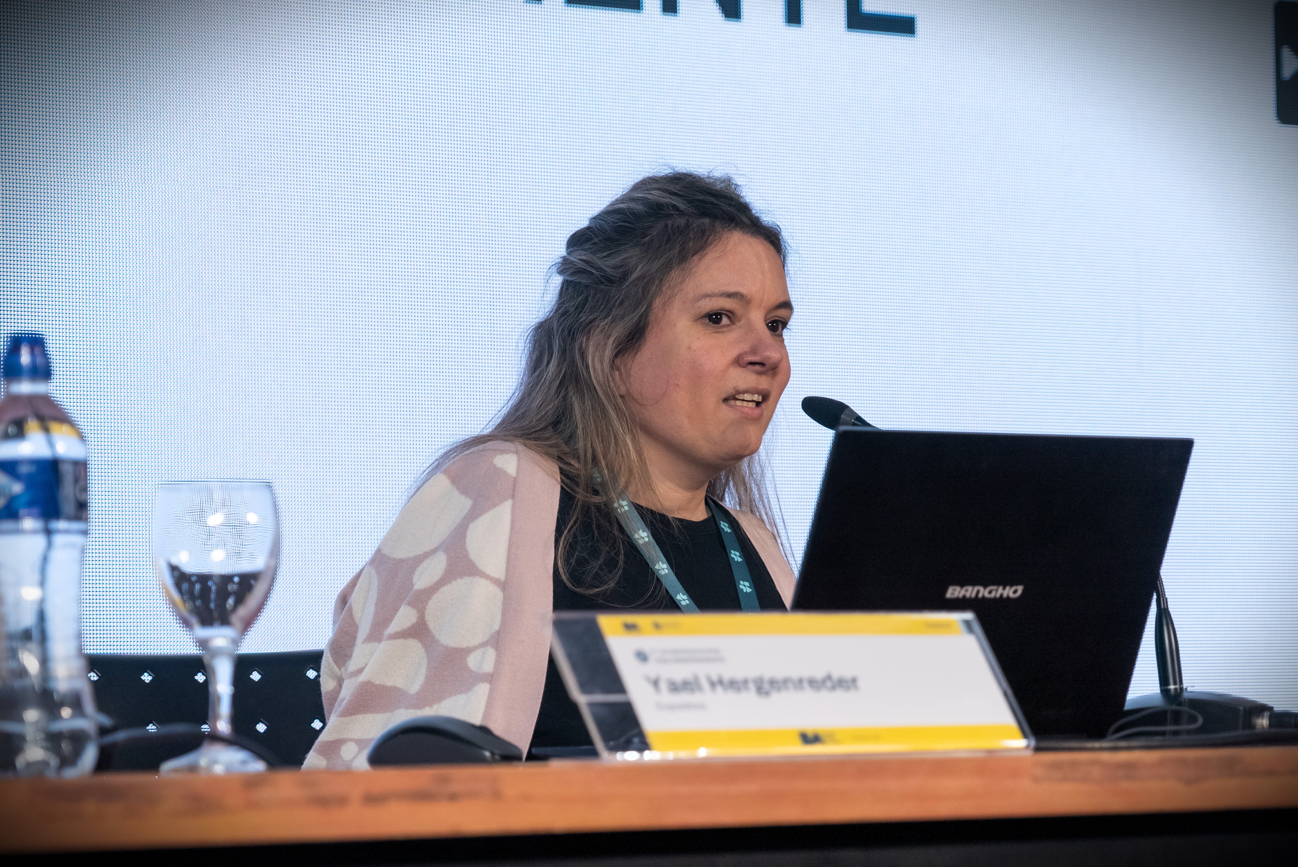 En la imagen se puede ver a Yael Hergenreder, que está participando en un evento.Está sentada en una mesa con un portátil frente a ella y una botella de agua al lado.