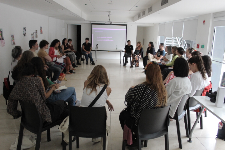 La imagen muestra un grupo de personas sentadas en círculo en una sala de reuniones. Hay aproximadamente 30 participantes, que parecen estar involucrados en una discusión o taller. En el centro, hay dos personas que están hablando o moderando la sesión. Detrás de ellos, hay una pantalla que probablemente muestra información relacionada con el encuentro.