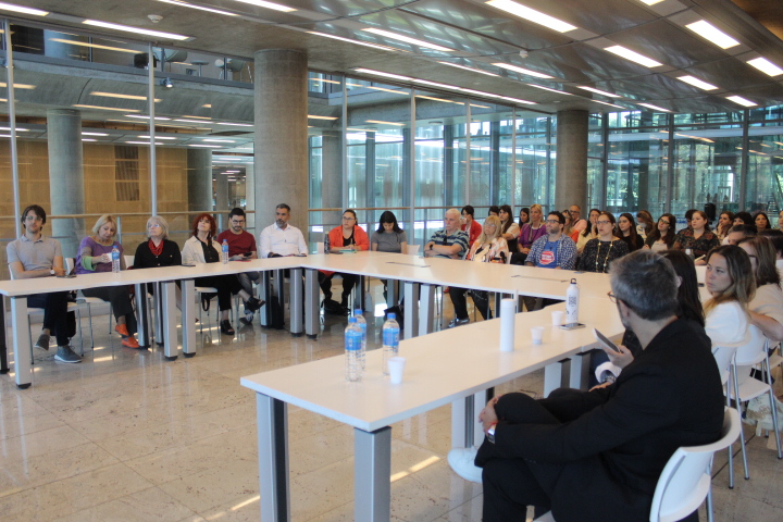 La imagen muestra una sala de reuniones moderna, donde un grupo de aproximadamente 30 personas está sentado alrededor de una larga mesa blanca en forma de U. El ambiente es luminoso, gracias a grandes ventanales que permiten la entrada de luz natural.  Los participantes son diversos en género y edad; algunos parecen estar atentos a un orador que se encuentra al frente de la mesa. 
