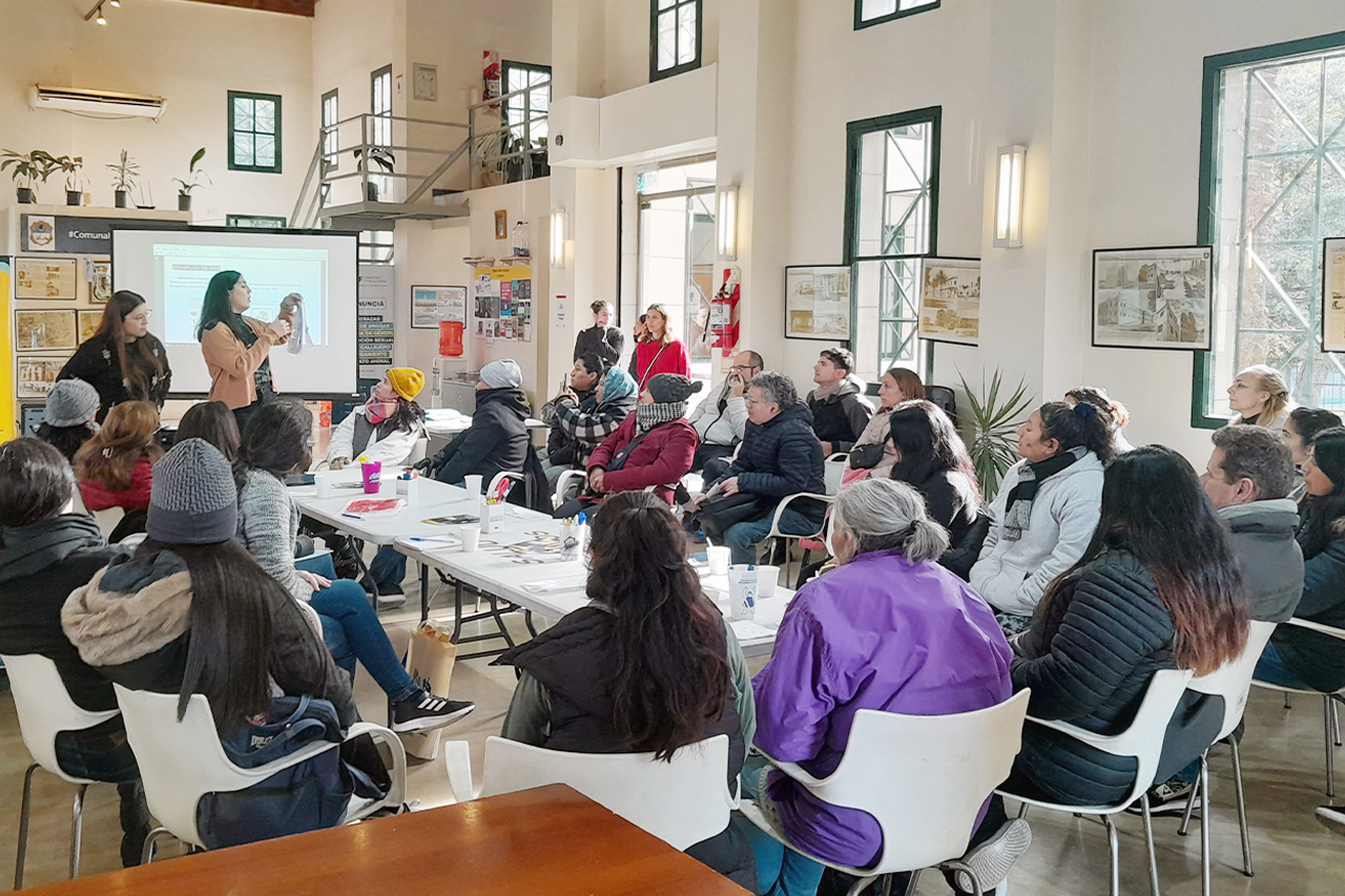Personas sentadas alrededor de una mesa larga participando de un taller en un espacio comunitario con una presentadora mostrando un material visual