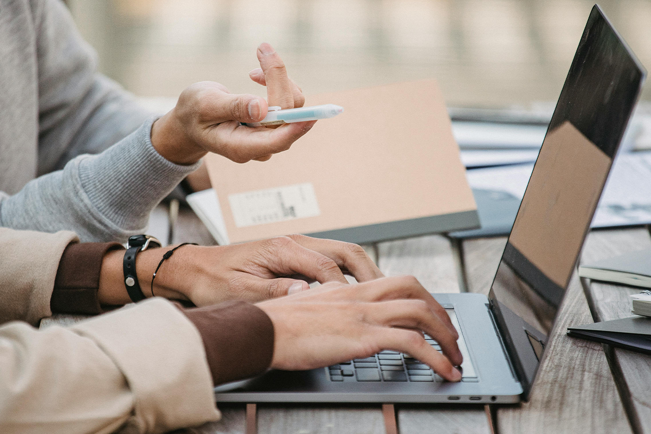 Dos personas colaborando en una computadora portátil al aire libre, con una de ellas escribiendo y la otra señalando con un bolígrafo sobre una mesa con cuadernos.