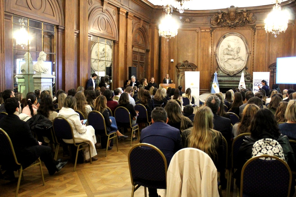 Salón de la Legislatura con público asistiendo a la presentación del Protocolo ProBono y una oradora en el escenario, rodeado de decoración de madera y lámparas colgantes.