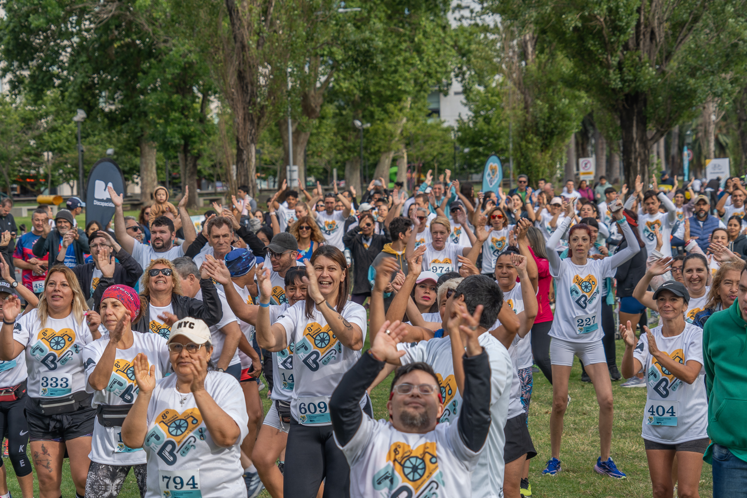 La imagen mustra a una gran multitud de personas participando de la carrera. Todos llevan la camiseta blanca con el logo diseño y numeros en el pecho de identiicacion. La atmofera es alegre y energetica, con muchas personas sonriendo y levantando la mano