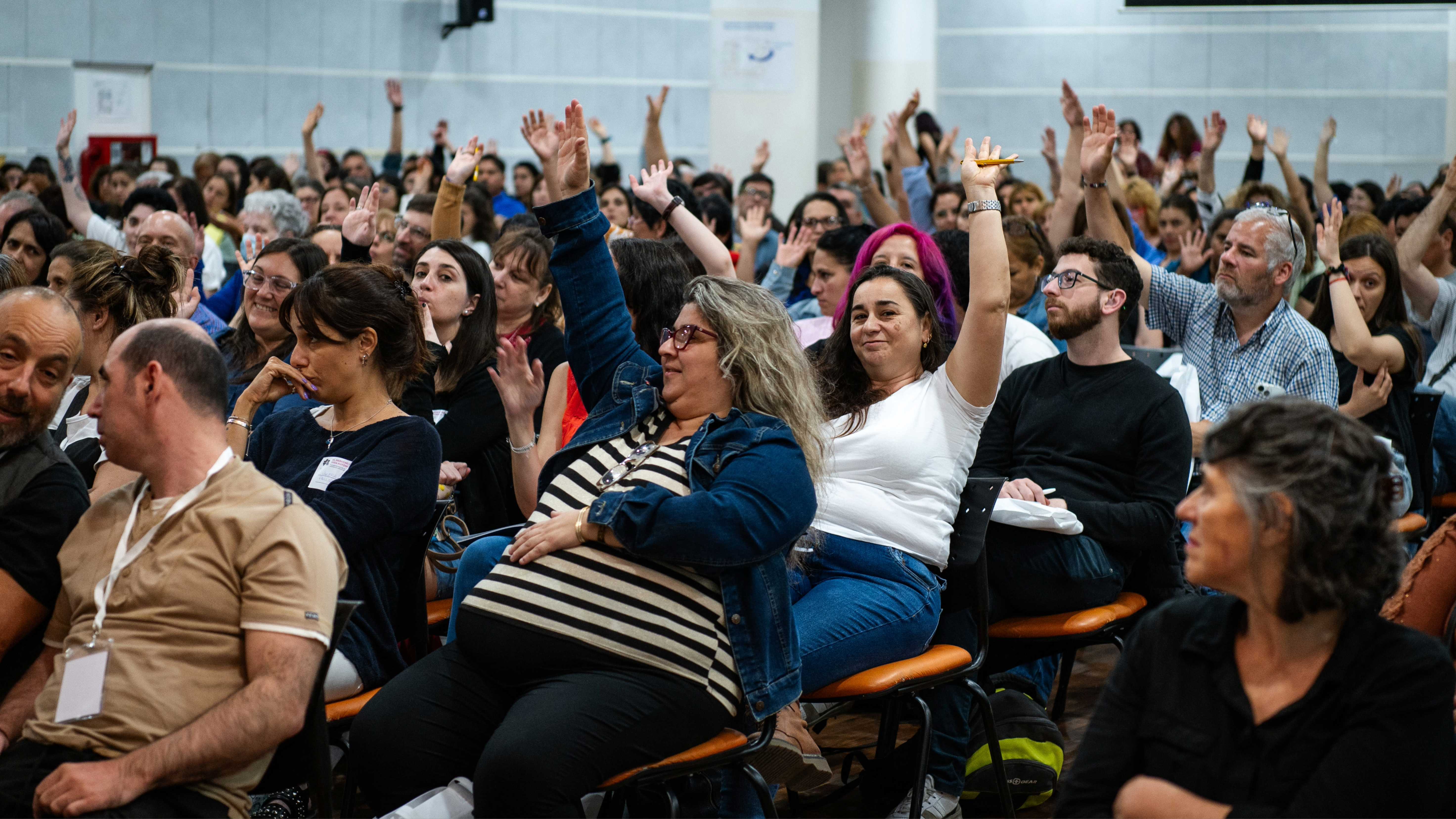 La imagen muestra a un gran grupo de personas presentes en la jornada. La mayoría de los asistentes están sentados y levantando la mano, lo que sugiere que están participando activamente en una discusión o votación.