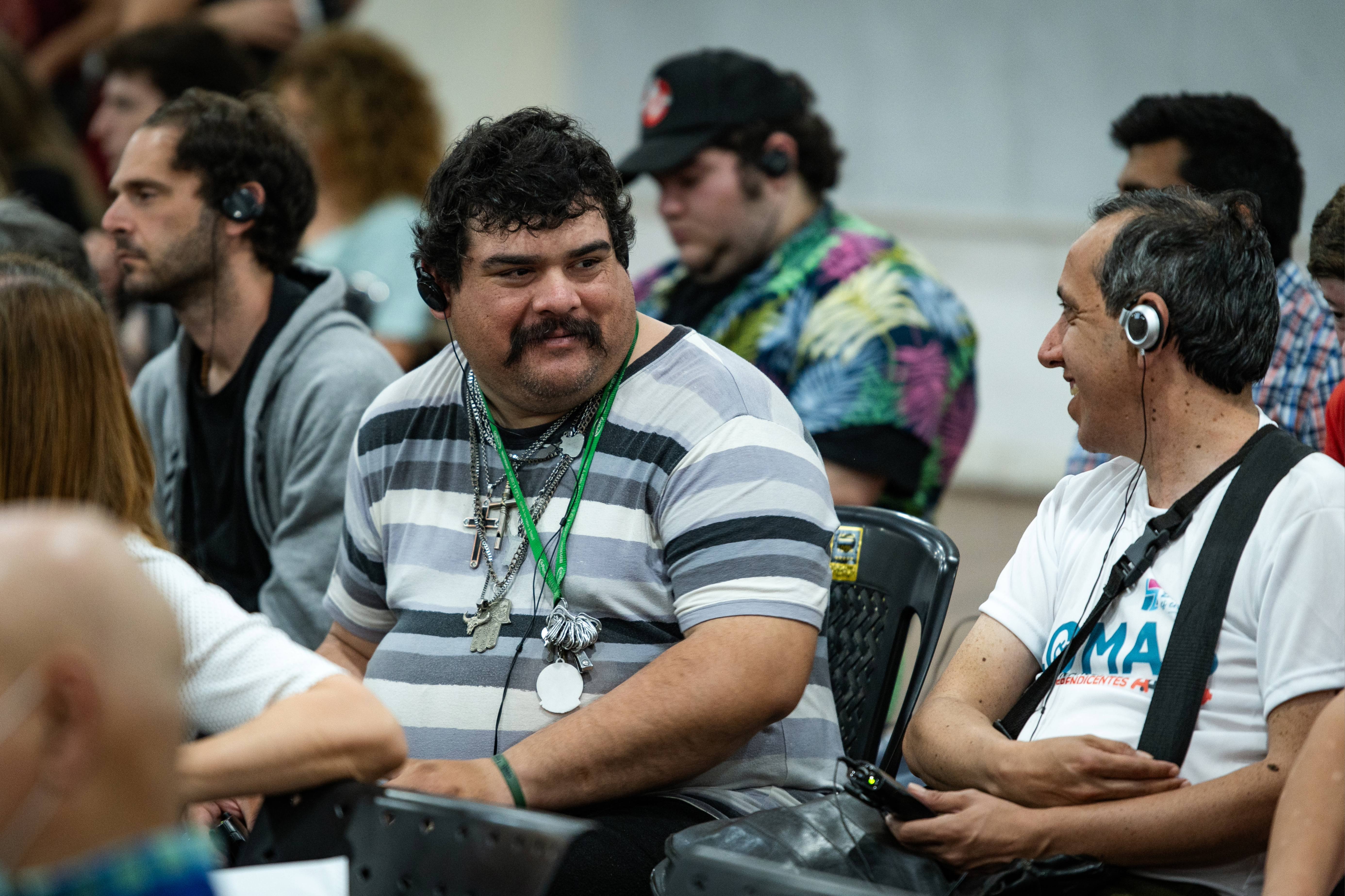 La imagen muestra a un grupo de personas sentadas en el evento. En el primer plano, hay un hombre de complexión robusta, con una camiseta de rayas y varios collares. Tiene una expresión amigable y está mirando a su lado, donde se encuentra otro hombre que lleva una camiseta blanca y auriculares. Este último parece estar sonriendo y conversando con el hombre de la camiseta de rayas.