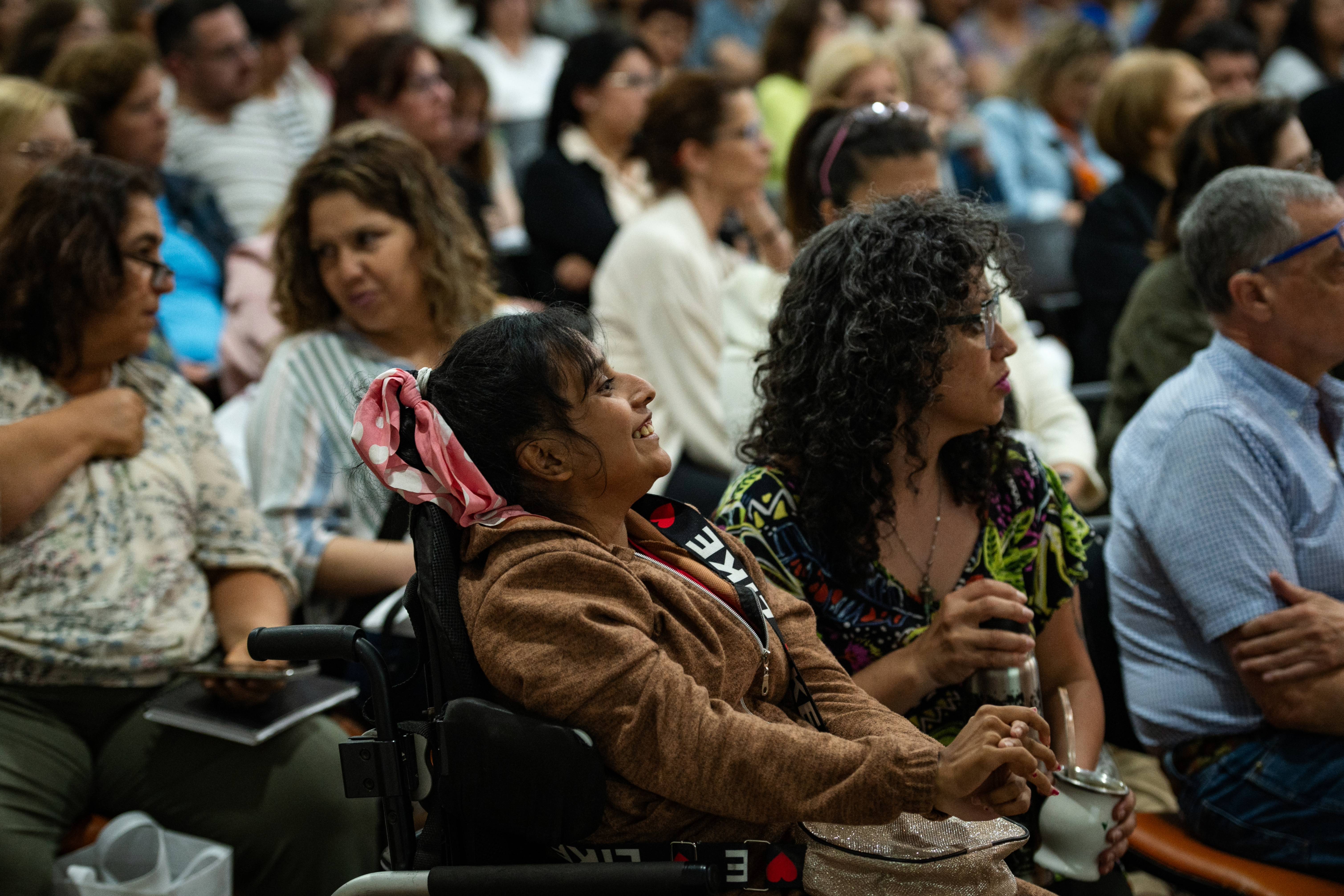 La imagen muestra a un grupo de personas participantes de una jornada. La mayoría de los asistentes son mujeres, y se pueden ver diferentes expresiones en sus rostros, que van desde la atención hasta la concentración. Algunas personas están usando dispositivos móviles, como teléfonos y tabletas, para tomar fotos o grabar.  En el primer plano, hay una mujer con un teléfono azul que parece estar capturando un momento del evento. Otra mujer, a su lado, sostiene una bolsa blanca con un diseño que parece estar relacionado con el evento. El ambiente es de interés y participación, con un fondo de personas que observan atentamente. La iluminación es suave, lo que sugiere un ambiente acogedor y profesional.