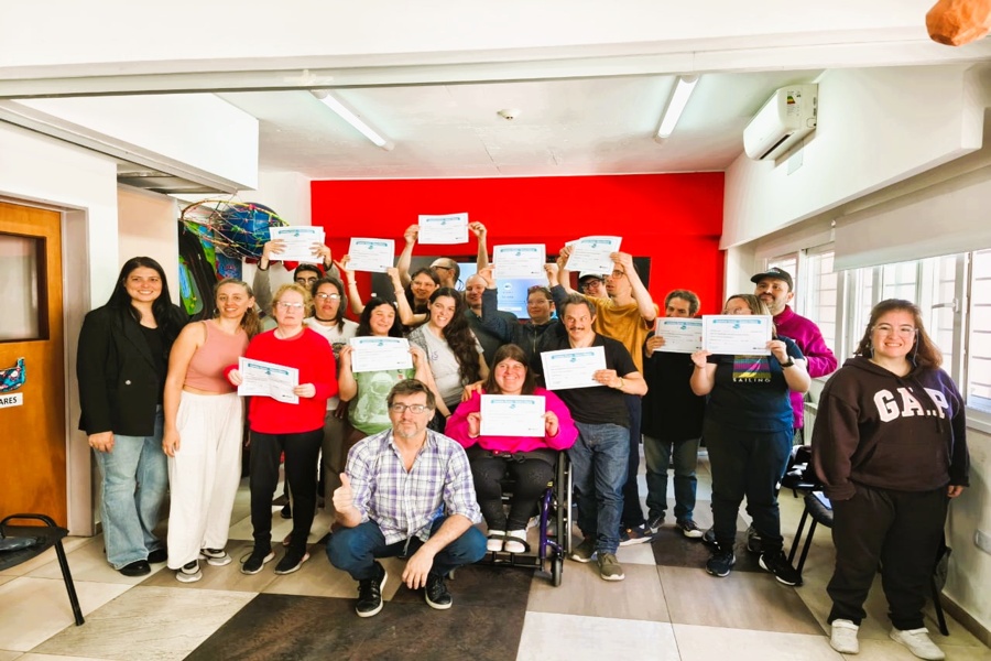 Participantes del taller y equipo de coordinación posando en un aula tras recibir sus certificados, con una pared roja de fondo y luz natural ingresando por las ventanas.