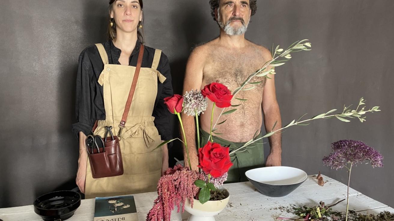 Dos personas posan detras de una mesa con flores e instrumentos para hacer arreglos florales