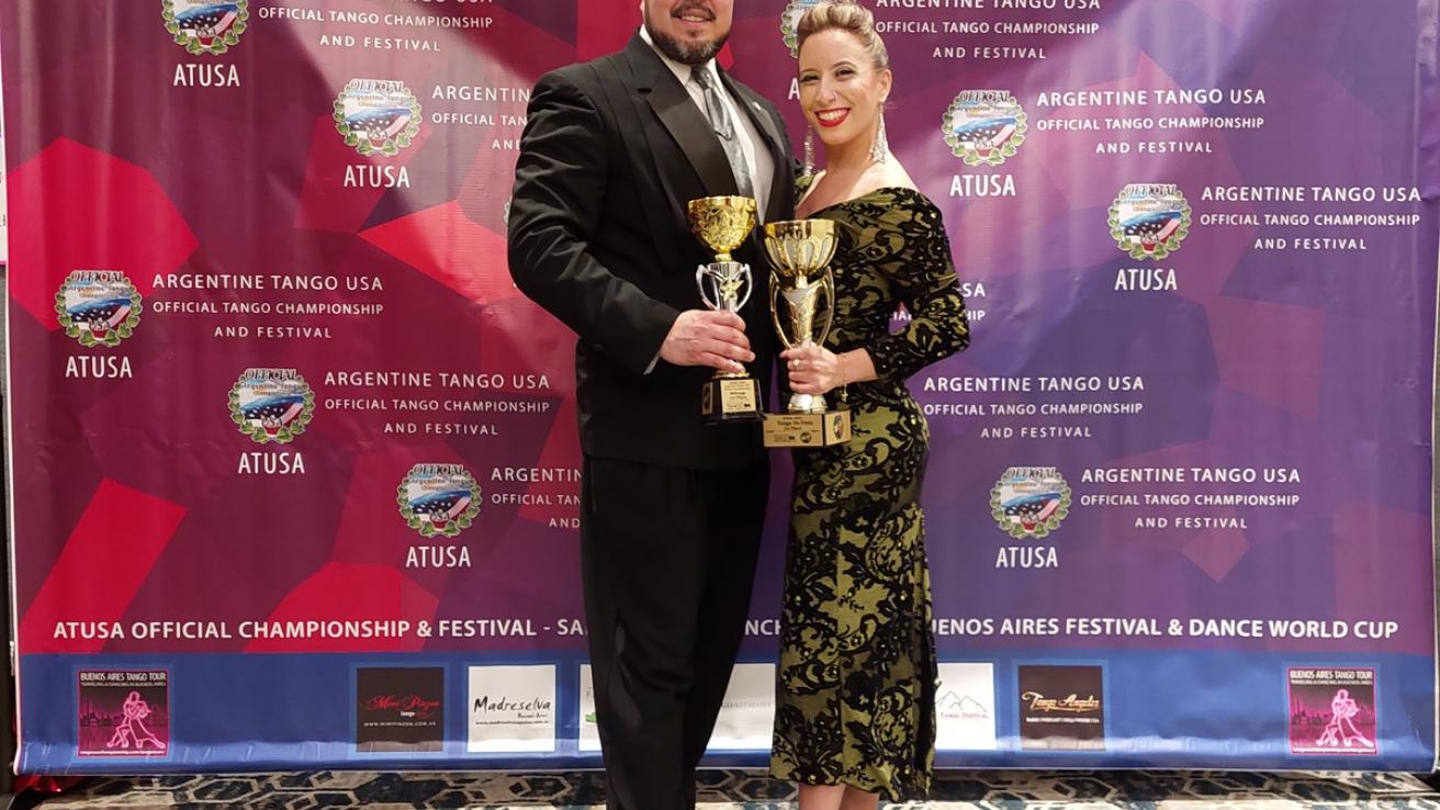 Pareja ganadora, mujer y hombre, de la categoria Tango de Pista, posan con sus dos trofeos delante del banner del evento Argentine Tango Usa