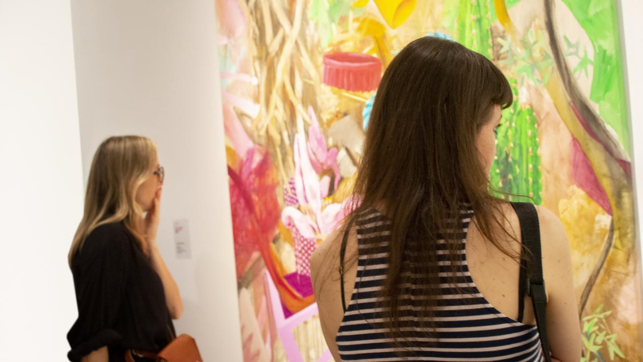 Imagen donde se observa un detalle de una sala de exposición con una obra de pintura y dos mujeres observándolapintura