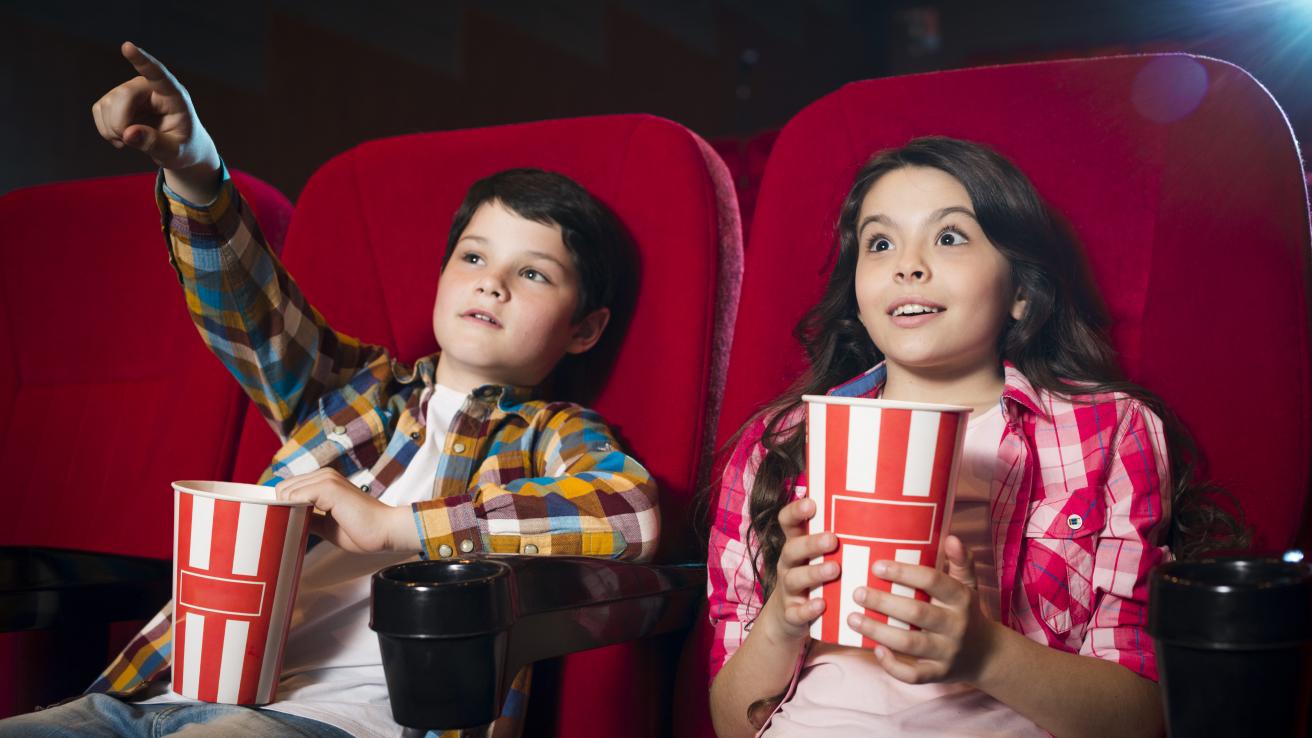 Foto de un niño y una niña sentados en una sala de cine con pochoclos en la mano y mirando una película