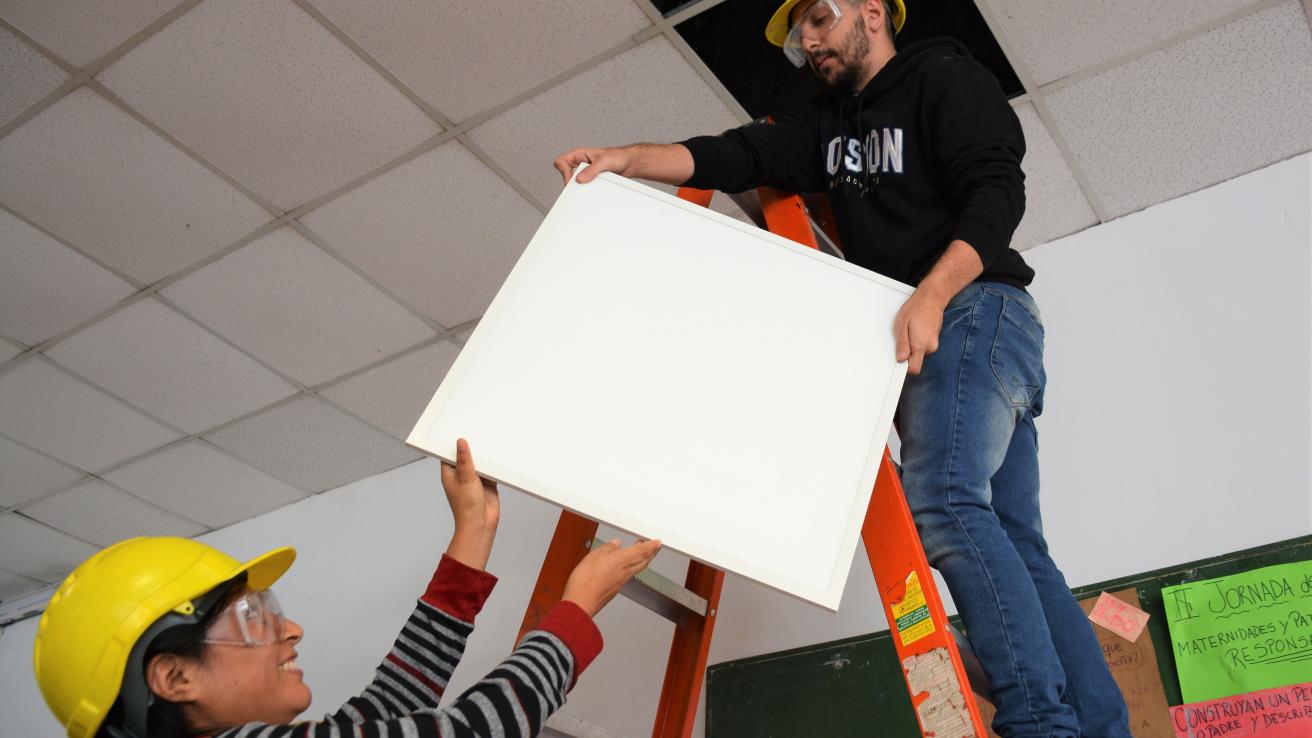 Dos hombres sostienen una pieza cuadrada blanca. Uno esta subido a una escalera