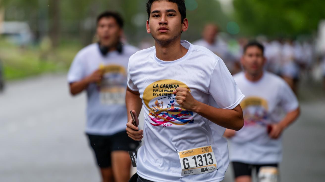 En la imagen se observa a un grupo de corredores participando en una carrera. En primer plano, un joven está corriendo con una expresión de concentración. Lleva una camiseta blanca con un diseño colorido que dice "LA CARRERA POR LA INCLUSIÓN EN LA CIUDAD". También tiene un número de corredor visible en su camiseta, "G 1313"