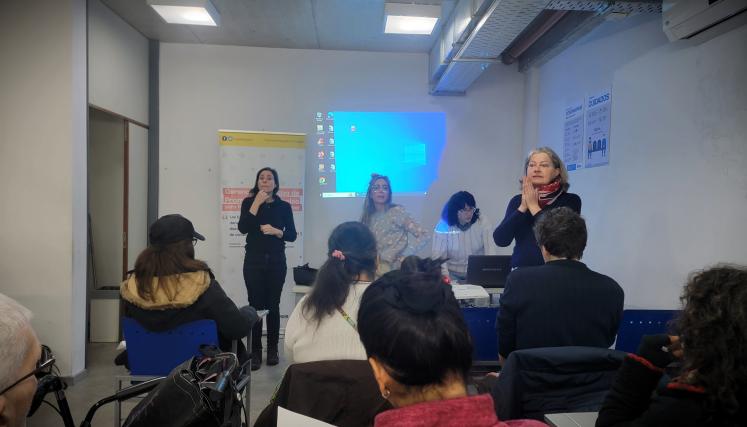 La imagen muestra un ambiente de presentación. Hay varias personas sentadas, algunas de espaldas, mirando hacia el frente donde se encuentran tres mujeres. Una de ellas está de pie, hablando. Otra mujer, también de pie, parece estar utilizando un dispositivo o computadora portátil. En el fondo, se puede ver una pantalla con un fondo azul, lo que sugiere que se está proyectando información. El ambiente parece ser de aprendizaje, con un enfoque en la interacción.