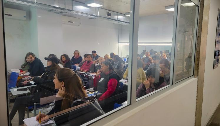 La imagen muestra un aula de clases con varias personas sentadas en escritorios. A través de un panel de vidrio, se pueden ver al menos veinte personas, la mayoría de ellas prestando atención hacia el frente. Algunas parecen estar tomando notas, mientras que otras están en diferentes posturas, como escuchando o reflexionando. El ambiente es de aprendizaje, y la iluminación es clara, lo que sugiere un espacio bien iluminado. Las paredes son de color claro, y hay un proyector o pantalla en la parte frontal de