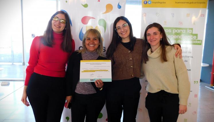 En la imagen se pueden ver a cuatro personas posando juntas en un ambiente interior. Tres son mujeres y están de pie, mientras que una está en el centro sosteniendo un certificado o diploma. Todas parecen estar sonriendo y celebrando un logro. Las mujeres están vestidas de manera casual, con suéteres y abrigos. Al fondo, se pueden ver dos carteles que parecen estar relacionados con un evento o una organización, con colores vivos y un diseño moderno. La iluminación es clara y el espacio parece ser amplio y a
