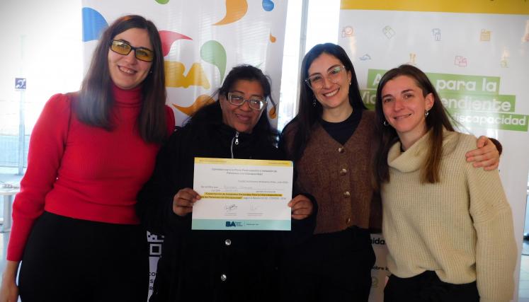 En la imagen se pueden ver a cuatro personas posando juntas en un ambiente interior. Tres son mujeres y están de pie, mientras que una está en el centro sosteniendo un certificado o diploma. Todas parecen estar sonriendo y celebrando un logro. Las mujeres están vestidas de manera casual, con suéteres y abrigos. Al fondo, se pueden ver dos carteles que parecen estar relacionados con un evento o una organización, con colores vivos y un diseño moderno. La iluminación es clara y el espacio parece ser amplio y a