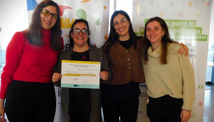En la imagen se pueden ver a cuatro personas posando juntas en un ambiente interior. Tres son mujeres y están de pie, mientras que una está en el centro sosteniendo un certificado o diploma. Todas parecen estar sonriendo y celebrando un logro. Las mujeres están vestidas de manera casual, con suéteres y abrigos. Al fondo, se pueden ver dos carteles que parecen estar relacionados con un evento o una organización, con colores vivos y un diseño moderno. La iluminación es clara y el espacio parece ser amplio y a