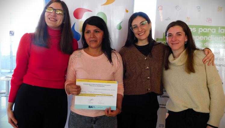En la imagen se pueden ver a cuatro personas posando juntas en un ambiente interior. Tres son mujeres y están de pie, mientras que una está en el centro sosteniendo un certificado o diploma. Todas parecen estar sonriendo y celebrando un logro. Las mujeres están vestidas de manera casual, con suéteres y abrigos. Al fondo, se pueden ver dos carteles que parecen estar relacionados con un evento o una organización, con colores vivos y un diseño moderno. La iluminación es clara y el espacio parece ser amplio y a