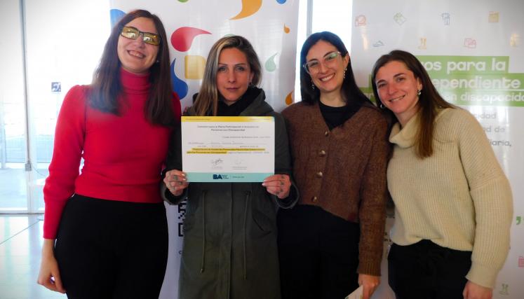 En la imagen se pueden ver a cuatro personas posando juntas en un ambiente interior. Tres son mujeres y están de pie, mientras que una está en el centro sosteniendo un certificado o diploma. Todas parecen estar sonriendo y celebrando un logro. Las mujeres están vestidas de manera casual, con suéteres y abrigos. Al fondo, se pueden ver dos carteles que parecen estar relacionados con un evento o una organización, con colores vivos y un diseño moderno. La iluminación es clara y el espacio parece ser amplio y a