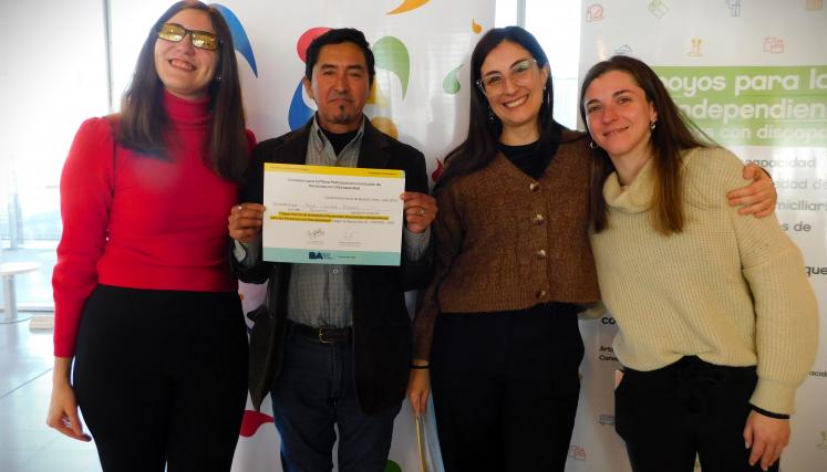 En la imagen se pueden ver a cuatro personas posando juntas en un ambiente interior. Tres son mujeres y están de pie, mientras que una está en el centro sosteniendo un certificado o diploma. Todas parecen estar sonriendo y celebrando un logro. Las mujeres están vestidas de manera casual, con suéteres y abrigos. Al fondo, se pueden ver dos carteles que parecen estar relacionados con un evento o una organización, con colores vivos y un diseño moderno. La iluminación es clara y el espacio parece ser amplio y a