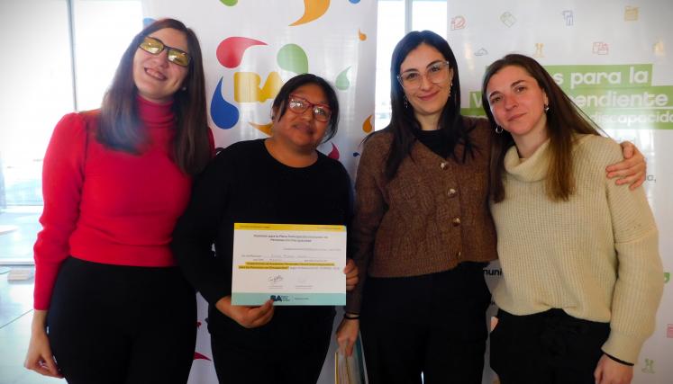 En la imagen se pueden ver a cuatro personas posando juntas en un ambiente interior. Tres son mujeres y están de pie, mientras que una está en el centro sosteniendo un certificado o diploma. Todas parecen estar sonriendo y celebrando un logro. Las mujeres están vestidas de manera casual, con suéteres y abrigos. Al fondo, se pueden ver dos carteles que parecen estar relacionados con un evento o una organización, con colores vivos y un diseño moderno. La iluminación es clara y el espacio parece ser amplio y a