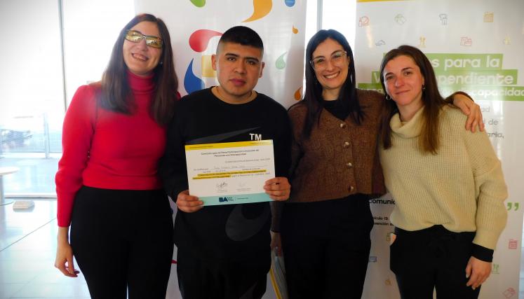 En la imagen se pueden ver a cuatro personas posando juntas en un ambiente interior. Tres son mujeres y están de pie, mientras que una está en el centro sosteniendo un certificado o diploma. Todas parecen estar sonriendo y celebrando un logro. Las mujeres están vestidas de manera casual, con suéteres y abrigos. Al fondo, se pueden ver dos carteles que parecen estar relacionados con un evento o una organización, con colores vivos y un diseño moderno. La iluminación es clara y el espacio parece ser amplio y a