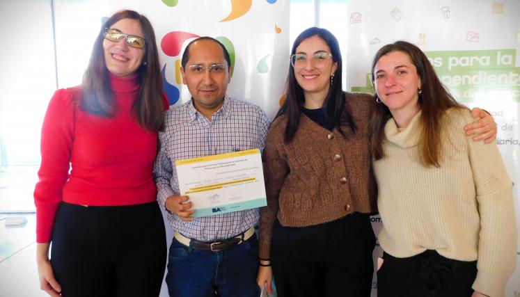 En la imagen se pueden ver a cuatro personas posando juntas en un ambiente interior. Tres son mujeres y están de pie, mientras que una está en el centro sosteniendo un certificado o diploma. Todas parecen estar sonriendo y celebrando un logro. Las mujeres están vestidas de manera casual, con suéteres y abrigos. Al fondo, se pueden ver dos carteles que parecen estar relacionados con un evento o una organización, con colores vivos y un diseño moderno. La iluminación es clara y el espacio parece ser amplio y a