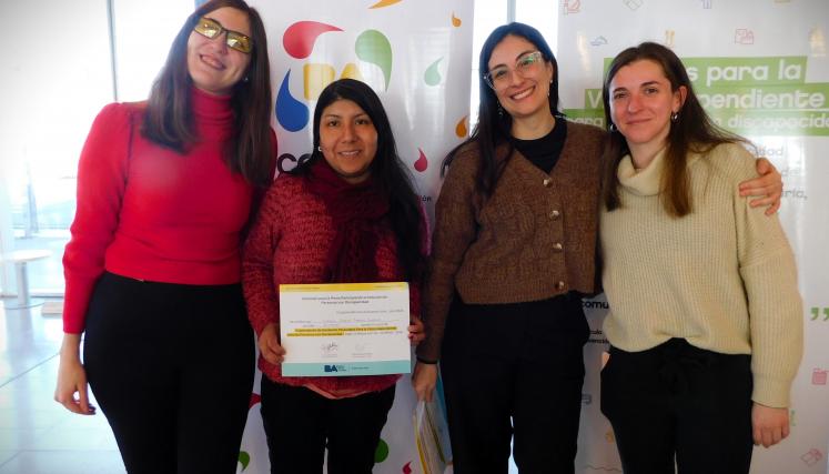 En la imagen se pueden ver a cuatro personas posando juntas en un ambiente interior. Tres son mujeres y están de pie, mientras que una está en el centro sosteniendo un certificado o diploma. Todas parecen estar sonriendo y celebrando un logro. Las mujeres están vestidas de manera casual, con suéteres y abrigos. Al fondo, se pueden ver dos carteles que parecen estar relacionados con un evento o una organización, con colores vivos y un diseño moderno. La iluminación es clara y el espacio parece ser amplio y a