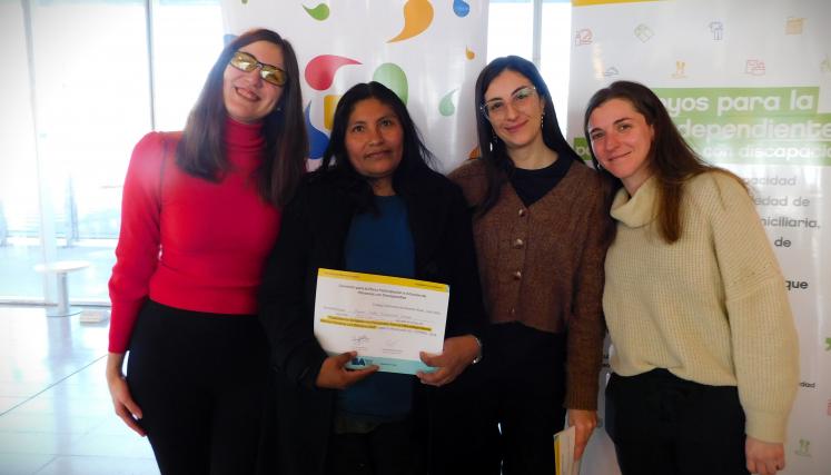 En la imagen se pueden ver a cuatro personas posando juntas en un ambiente interior. Tres son mujeres y están de pie, mientras que una está en el centro sosteniendo un certificado o diploma. Todas parecen estar sonriendo y celebrando un logro. Las mujeres están vestidas de manera casual, con suéteres y abrigos. Al fondo, se pueden ver dos carteles que parecen estar relacionados con un evento o una organización, con colores vivos y un diseño moderno. La iluminación es clara y el espacio parece ser amplio y a