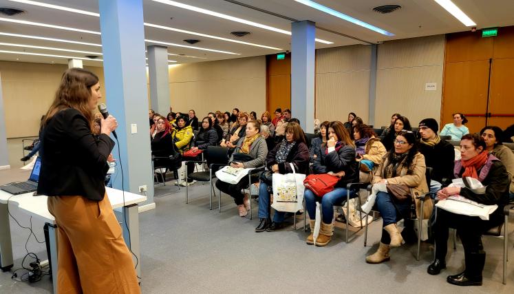 La imagen muestra a Victoria Belotti de pie, hablando frente a un micrófono en un ambiente de presentación. Ella tiene el cabello largo y oscuro, y viste una blusa negra con pantalones de color claro. Su expresión es amigable y entusiasta, lo que sugiere que está compartiendo información o motivando a la audiencia. 