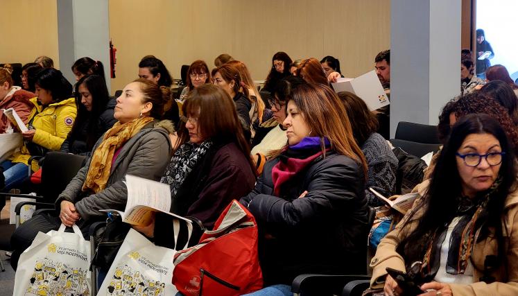 La imagen muestra a un grupo de mujeres sentadas en un auditorio, concentradas en la lectura de documentos que tienen en sus manos. La mayoría de ellas viste ropa de abrigo, como chaquetas y bufandas, lo que sugiere que el ambiente es fresco. Algunas tienen bolsas a su lado, con materiales relacionados con el evento. Las expresiones en sus rostros son serias y atentas, indicando que están involucradas en la capacitación o presentación. En el fondo, se pueden ver más asistentes, lo que sugiere una buena part