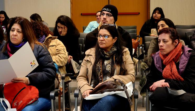 La imagen muestra a un grupo de mujeres sentadas en un auditorio, concentradas en la lectura de documentos que tienen en sus manos. La mayoría de ellas viste ropa de abrigo, como chaquetas y bufandas, lo que sugiere que el ambiente es fresco. Algunas tienen bolsas a su lado, con materiales relacionados con el evento. Las expresiones en sus rostros son serias y atentas, indicando que están involucradas en la capacitación o presentación. En el fondo, se pueden ver más asistentes, lo que sugiere una buena part