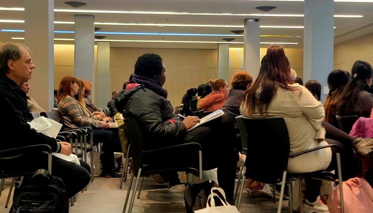 La imagen muestra a un grupo de personas sentadas en un auditorio, prestando atención a una presentación. La mayoría son mujeres, con una variedad de estilos de cabello y vestimenta. Algunas llevan chaquetas y bufandas, lo que sugiere que el ambiente es fresco. En el primer plano, se puede ver a un hombre con cabello castaño claro, que parece estar concentrado en lo que está sucediendo. A su lado, hay una mujer con un abrigo oscuro y una bufanda, que sostiene un cuaderno y un bolígrafo, indicando que está t