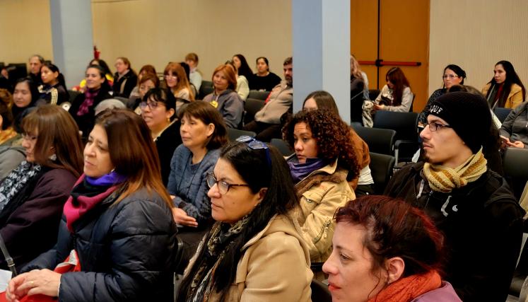 La imagen muestra a un grupo de personas sentadas en un auditorio, concentradas en una presentación. La mayoría son mujeres, con una variedad de estilos de cabello y vestimenta. Algunas llevan chaquetas y bufandas, lo que sugiere que el ambiente es fresco. En el primer plano, se puede ver a varias mujeres con expresiones serias y atentas, algunas tomando notas. También hay un hombre al fondo, con un gorro negro y una bufanda de rayas, que parece estar escuchando con interés. La sala tiene un diseño moderno 