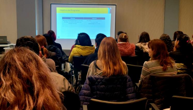 La imagen muestra a un grupo de personas sentadas en un auditorio, enfocadas en una presentación que se proyecta en una pantalla al frente. La mayoría de los asistentes son mujeres, con diferentes estilos de cabello y vestimenta. Algunas llevan chaquetas y abrigos, lo que sugiere que el ambiente es fresco. En la pantalla se pueden ver los "Objetivos del Programa", con dos columnas que indican los beneficiarios directos e indirectos del programa, lo que sugiere que se trata de una capacitación o presentación