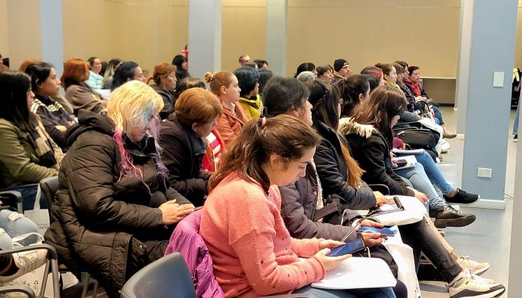 La imagen muestra a un grupo de personas sentadas en un auditorio, concentradas en una presentación. La mayoría son mujeres, con una variedad de estilos de cabello y vestimenta. Algunas llevan chaquetas y bufandas, lo que sugiere que el ambiente es fresco. En el primer plano, se puede ver a varias mujeres con expresiones serias y atentas, algunas tomando notas. También hay un hombre al fondo, con un gorro negro y una bufanda de rayas, que parece estar escuchando con interés. La sala tiene un diseño moderno 