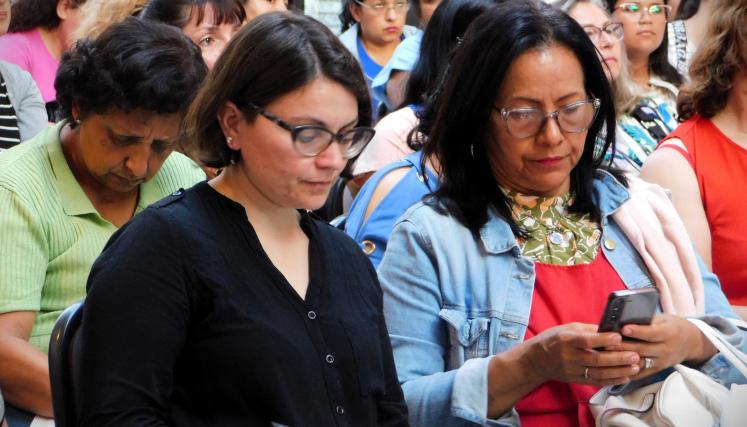 Imagen en la que destacanb dos mujeres. Una de ellas parece tomar apuntes y la otra mira su teléfono celular