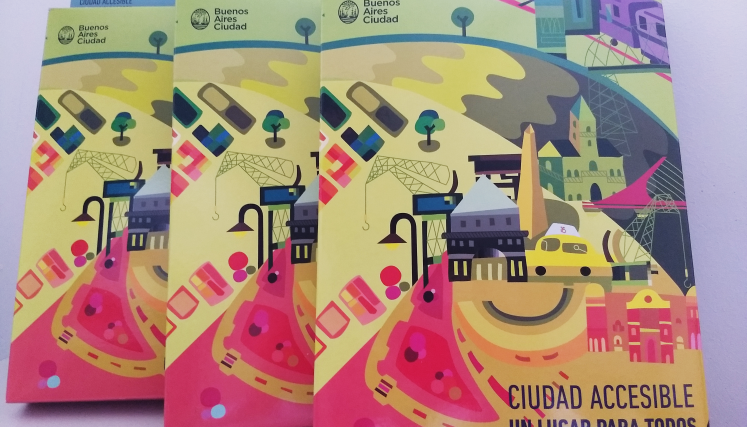 Tres folletos titulados 'Ciudad Accesible: Un lugar para todos' con una ilustración colorida que muestra elementos característicos de Buenos Aires, como el Obelisco, edificios históricos, un taxi, y grúas. Los folletos tienen un diseño en tonos amarillos, rosas, verdes y azules