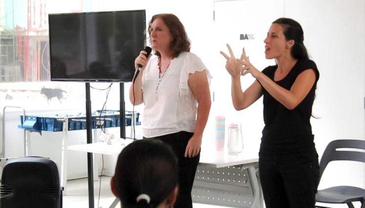 Mujer dando una charla con micrófono junto a intérprete de Lengua de Señas Argentina en una sala con personas presentes