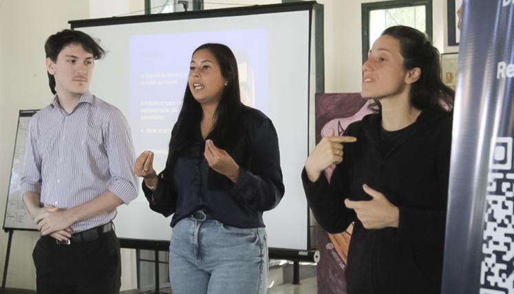 Tres personas de pie frente a una presentación; una mujer habla al público mientras otra interpreta en Lengua de Señas Argentina
