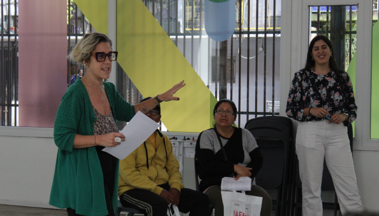 Una oradora de pie con una hoja en mano al lado de dos mujeres sentadas y otra parada